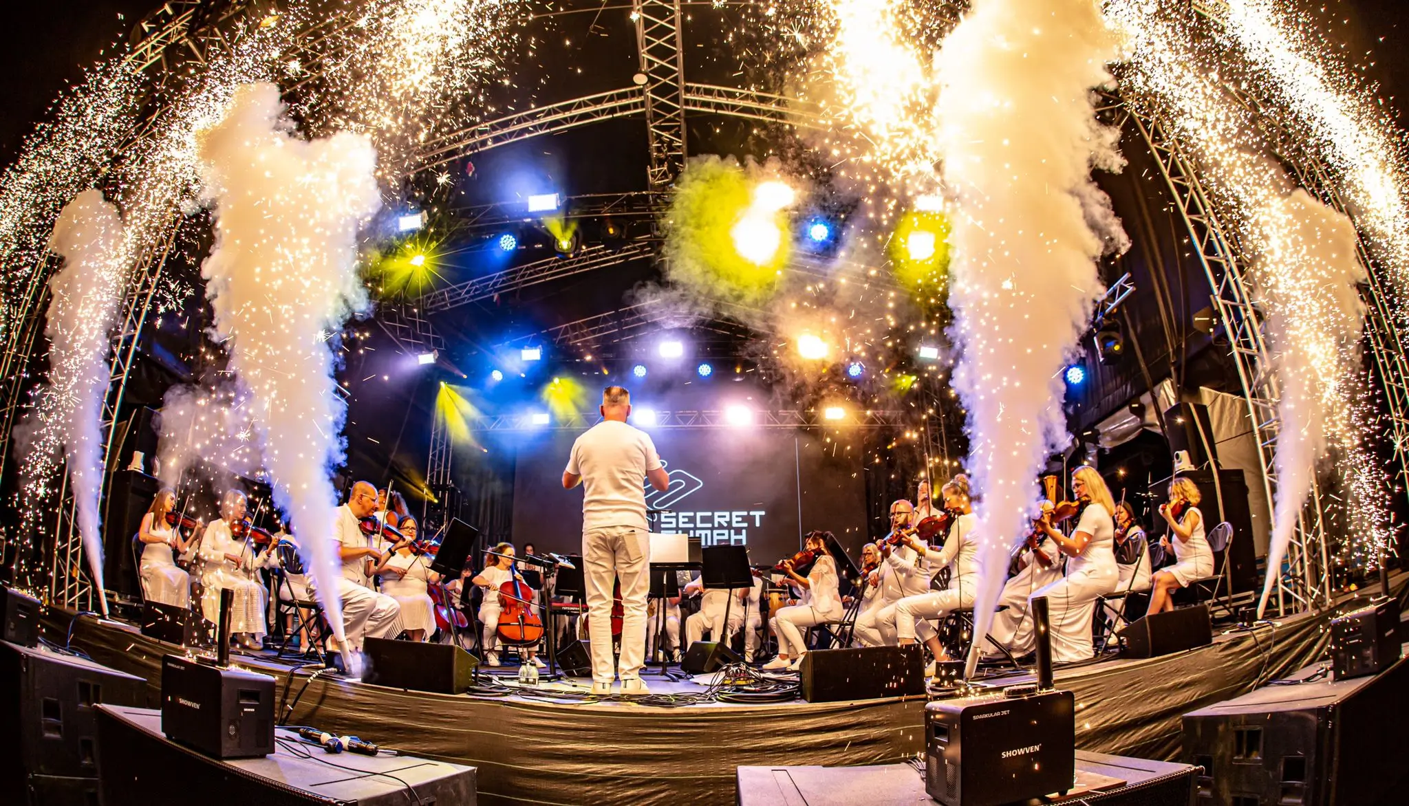 Orchestra performance on stage with pyrotechnics, smoke, and bright lights.